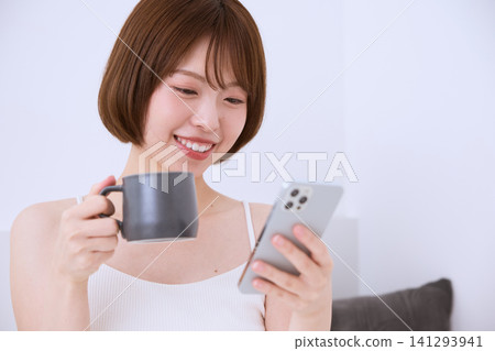 一位年輕女子麵帶微笑,一邊喝咖啡一邊看著她的智慧型手機。 一位年輕女子麵帶微笑,一邊喝咖啡一邊看著她的智慧型手機。 141293941