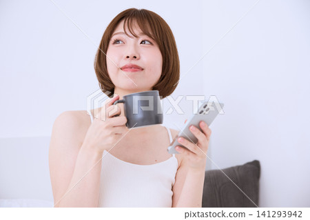 一位年輕女子正悠閒地玩著智慧型手機,手裡拿著咖啡杯。 一位年輕女子正悠閒地玩著智慧型手機,手裡拿著咖啡杯。 141293942