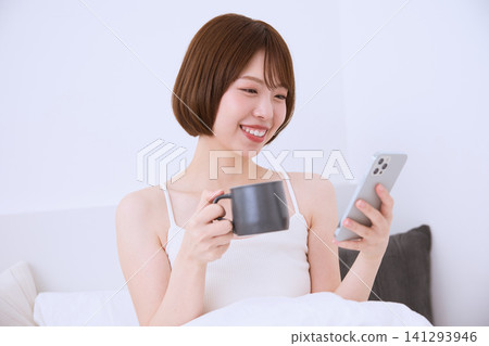 A young woman with a smile is drinking coffee while looking at her smartphone on her bed. A young woman with a smile is drinking coffee while looking at her smartphone on her bed. 141293946