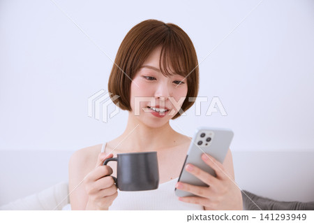 A young woman with short hair drinking coffee while looking at her smartphone. A young woman with short hair drinking coffee while looking at her smartphone. 141293949