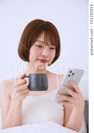 A relaxed young woman in bed, holding a coffee cup and a smartphone. A relaxed young woman in bed, holding a coffee cup and a smartphone. 141293954