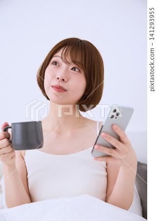 A young woman on a bed, holding a smartphone and a coffee cup. 141293955