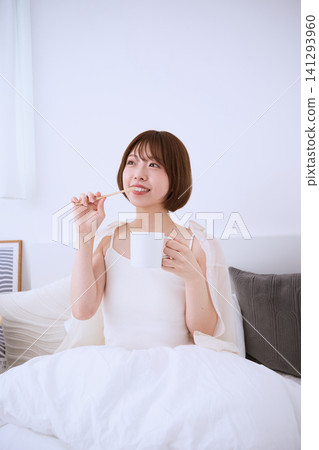清晨,一位年輕女子坐在床上,手裡拿著牙刷和杯子。 清晨,一位年輕女子坐在床上,手裡拿著牙刷和杯子。 141293960