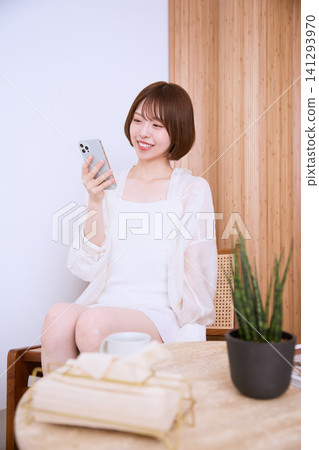 一位年輕女子坐在沙發上,看著她的智慧型手機,面帶微笑。 一位年輕女子坐在沙發上,看著她的智慧型手機,面帶微笑。 141293970