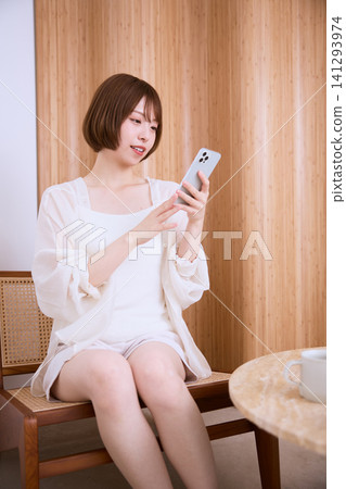 一位身穿白色衣服的年輕女子坐在椅子上操作她的智慧型手機。 一位身穿白色衣服的年輕女子坐在椅子上操作她的智慧型手機。 141293974