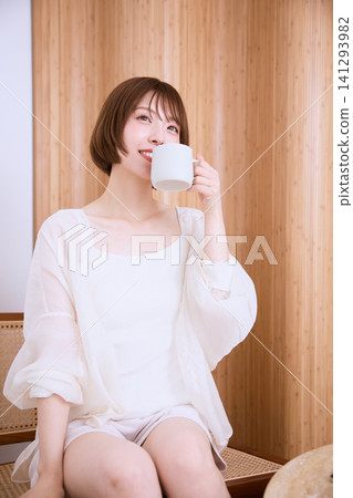 A young woman in a white dress is sitting in a chair and holding a coffee cup. A young woman in a white dress is sitting in a chair and holding a coffee cup. 141293982