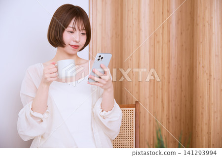一位留著波波頭的年輕女子一邊喝咖啡一邊看著她的智慧型手機。 一位留著波波頭的年輕女子一邊喝咖啡一邊看著她的智慧型手機。 141293994