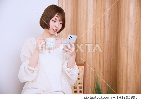 一位留著短髮的年輕女子一邊喝咖啡一邊看著她的智慧型手機。 一位留著短髮的年輕女子一邊喝咖啡一邊看著她的智慧型手機。 141293995