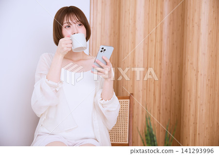 A relaxed young woman drinking coffee while looking at her smartphone. 141293996