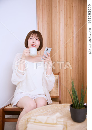 一位面帶微笑的年輕女子在客廳放鬆地玩著智慧型手機,手裡拿著一杯咖啡。 一位面帶微笑的年輕女子在客廳放鬆地玩著智慧型手機,手裡拿著一杯咖啡。 141293999
