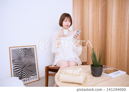 一位年輕女子在家一邊喝咖啡一邊看手機。 一位年輕女子在家一邊喝咖啡一邊看手機。 141294000