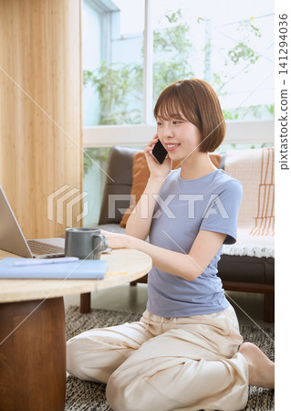 一位年輕女子麵帶微笑,在客廳用智慧型手機通話。 一位年輕女子麵帶微笑,在客廳用智慧型手機通話。 141294036