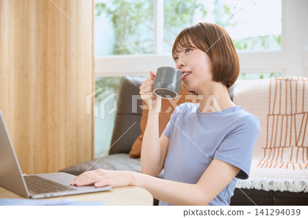 一位年輕女子在客廳裡一邊喝咖啡一邊使用筆記型電腦。 一位年輕女子在客廳裡一邊喝咖啡一邊使用筆記型電腦。 141294039