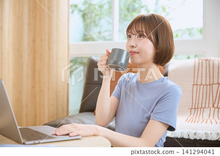 一位年輕女子在客廳裡一邊喝咖啡一邊使用筆記型電腦。 141294041