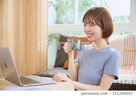 一位面帶微笑的年輕女子在客廳裡一邊喝咖啡一邊使用筆記型電腦。 一位面帶微笑的年輕女子在客廳裡一邊喝咖啡一邊使用筆記型電腦。 141294042