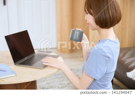 一位年輕女子在客廳裡一邊喝咖啡一邊使用筆記型電腦。 一位年輕女子在客廳裡一邊喝咖啡一邊使用筆記型電腦。 141294047