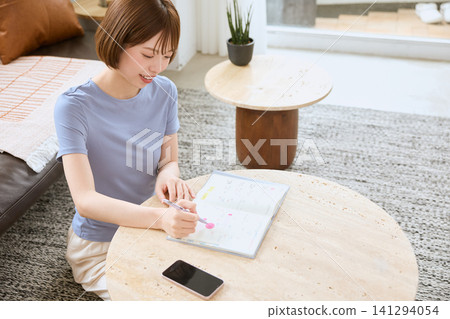 A young woman with a smile writing in her schedule book in the living room. A young woman with a smile writing in her schedule book in the living room. 141294054