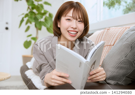 一位年輕女子麵帶微笑,坐在沙發上看書。 一位年輕女子麵帶微笑,坐在沙發上看書。 141294080