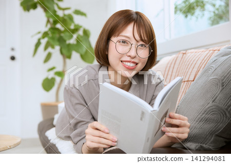 一位戴著眼鏡的年輕女子正放鬆地躺在沙發上看書。 一位戴著眼鏡的年輕女子正放鬆地躺在沙發上看書。 141294081