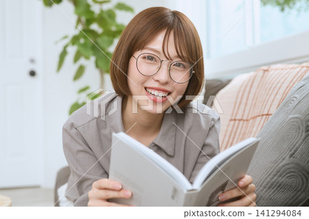 A young woman with glasses smiles while reading a book on a sofa. A young woman with glasses smiles while reading a book on a sofa. 141294084