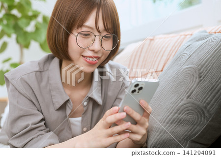 A young woman wearing glasses is smiling and looking at her smartphone on a sofa. A young woman wearing glasses is smiling and looking at her smartphone on a sofa. 141294091