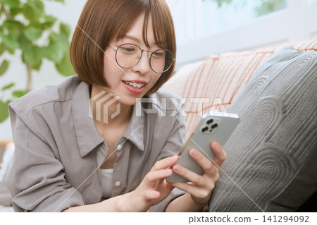 一位戴著眼鏡的年輕女子坐在沙發上,一邊看著她的智慧型手機一邊微笑。 一位戴著眼鏡的年輕女子坐在沙發上,一邊看著她的智慧型手機一邊微笑。 141294092