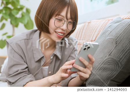 一位戴著眼鏡的年輕女子坐在沙發上,一邊看著她的智慧型手機一邊微笑。 一位戴著眼鏡的年輕女子坐在沙發上,一邊看著她的智慧型手機一邊微笑。 141294093