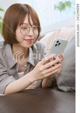 一位戴眼鏡的年輕女子正坐在沙發上看她的智慧型手機。 一位戴眼鏡的年輕女子正坐在沙發上看她的智慧型手機。 141294094