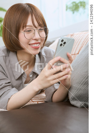 一位戴著眼鏡的年輕女子坐在沙發上,一邊看著她的智慧型手機一邊微笑。 一位戴著眼鏡的年輕女子坐在沙發上,一邊看著她的智慧型手機一邊微笑。 141294096