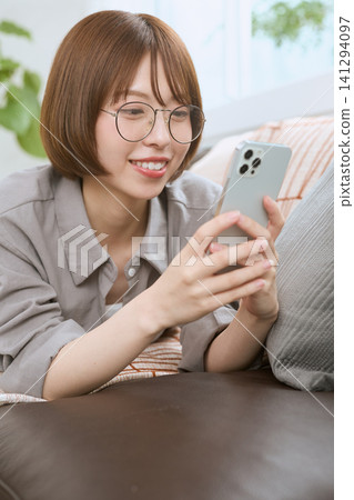 一位戴著眼鏡的年輕女子坐在沙發上,一邊看著她的智慧型手機一邊微笑。 一位戴著眼鏡的年輕女子坐在沙發上,一邊看著她的智慧型手機一邊微笑。 141294097