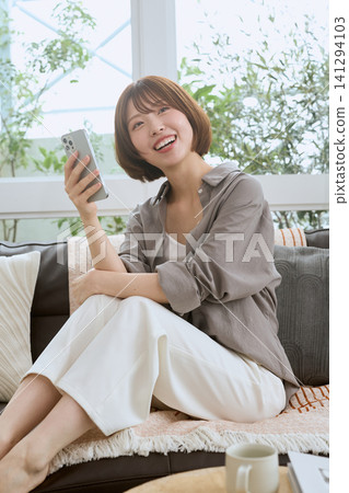 一位年輕女子坐在沙發上,看著她的智慧型手機,面帶微笑。 一位年輕女子坐在沙發上,看著她的智慧型手機,面帶微笑。 141294103