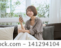 一位年輕女子坐在客廳沙發上,一邊看著她的智慧型手機一邊微笑。 一位年輕女子坐在客廳沙發上,一邊看著她的智慧型手機一邊微笑。 141294104