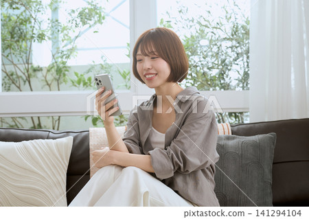 一位年輕女子坐在客廳沙發上,一邊看著她的智慧型手機一邊微笑。 一位年輕女子坐在客廳沙發上,一邊看著她的智慧型手機一邊微笑。 141294104