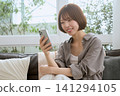 一位年輕女子坐在沙發上,看著她的智慧型手機,面帶微笑。 一位年輕女子坐在沙發上,看著她的智慧型手機,面帶微笑。 141294105