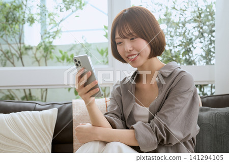 A young woman sitting on a sofa, looking at her smartphone and smiling. A young woman sitting on a sofa, looking at her smartphone and smiling. 141294105