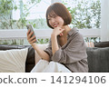 一位年輕女子坐在沙發上,一邊看著她的智慧型手機一邊微笑。 一位年輕女子坐在沙發上,一邊看著她的智慧型手機一邊微笑。 141294106