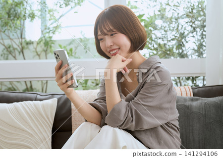 一位年輕女子坐在沙發上,一邊看著她的智慧型手機一邊微笑。 一位年輕女子坐在沙發上,一邊看著她的智慧型手機一邊微笑。 141294106