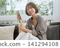 一位留著波波頭的年輕女子坐在沙發上,面帶微笑,手裡拿著她的智慧型手機。 一位留著波波頭的年輕女子坐在沙發上,面帶微笑,手裡拿著她的智慧型手機。 141294108