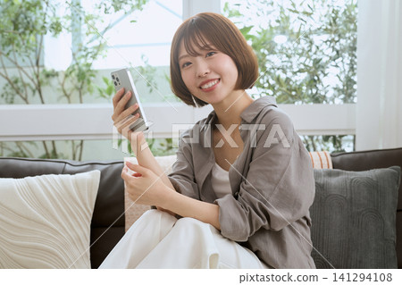 一位留著波波頭的年輕女子坐在沙發上,面帶微笑,手裡拿著她的智慧型手機。 一位留著波波頭的年輕女子坐在沙發上,面帶微笑,手裡拿著她的智慧型手機。 141294108