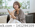一位年輕女子坐在沙發上,拿著智慧型手機,面帶微笑。 一位年輕女子坐在沙發上,拿著智慧型手機,面帶微笑。 141294109
