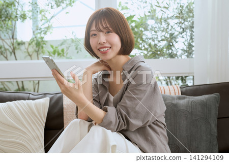 一位年輕女子坐在沙發上，拿著智慧型手機，面帶微笑。 141294109