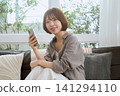 一位年輕女子坐在客廳沙發上,一邊拿著智慧型手機一邊微笑。 一位年輕女子坐在客廳沙發上,一邊拿著智慧型手機一邊微笑。 141294110