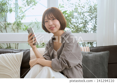 一位年輕女子坐在客廳沙發上,一邊拿著智慧型手機一邊微笑。 一位年輕女子坐在客廳沙發上,一邊拿著智慧型手機一邊微笑。 141294110
