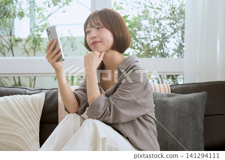 一位年輕女子坐在沙發上,一邊看著她的智慧型手機一邊微笑。 一位年輕女子坐在沙發上,一邊看著她的智慧型手機一邊微笑。 141294111