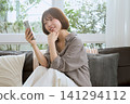 一位年輕女子坐在沙發上,看著她的智慧型手機,面帶微笑。 一位年輕女子坐在沙發上,看著她的智慧型手機,面帶微笑。 141294112