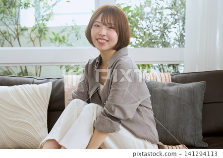 A young woman with short hair sits on a sofa and smiles. A young woman with short hair sits on a sofa and smiles. 141294113