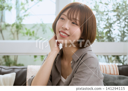 A young woman with a short bob haircut sits on a cafe sofa, smiling and relaxing. A young woman with a short bob haircut sits on a cafe sofa, smiling and relaxing. 141294115