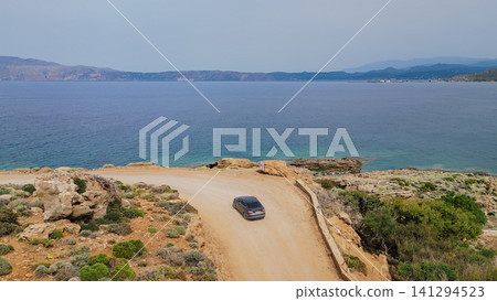 Horizontal high angle view of a gray car navigating a tight bend on a dusty coastal dirt road near Kissamos on Crete island in Greece with the deep blue sea behind 141294523