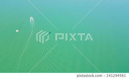 Horizontal aerial view of a speed boat towing a white parasail with a distinct dark shadow cast on the emerald sea surface during a sunny summer vacation 141294561