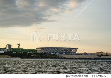 Volgograd, Russia - 05.25.2023: View of Volgograd Arena stadium on the banks of the Volga River with The Motherland Calls monument in the background. Evening cityscape with dramatic sky and calm water 141296078